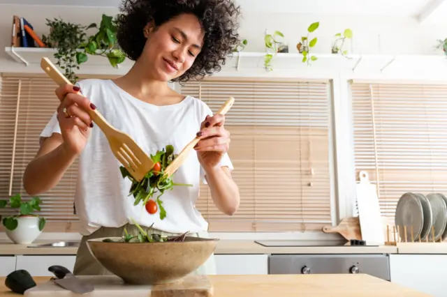 Mujer preparando una ensalada fresca en una cocina moderna, usando utensilios de madera para mezclar hojas verdes y tomates en un bol grande.