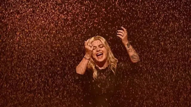 Plava žena peva sa rukama podignutim u vis, dok oko nje pada zlatni pesak