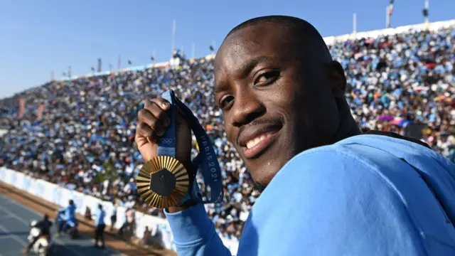 Letsile Tebogo regarde en arrière, brandit sa médaille d'or olympique de Paris 2024 et sourit à la caméra, tandis qu'une tribune remplie de milliers de supporters est visible au loin derrière lui.