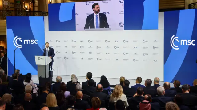 JD Vance, en un traje oscuro, se dirige a un público desde un podio, flanqueado por dos letreros azules que leen: "MSC" (Conferencia de Seguridad de Múnich)