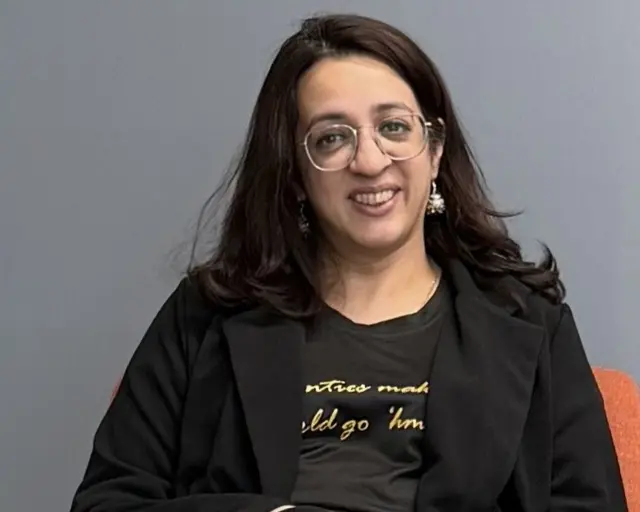 A woman with long brown hair and glasses smiles in a black top and blazer, in front of a grey background
