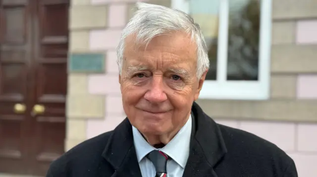 Un hombre que viste camisa y corbata con un abrigo encima está parado frente a un edificio de oficinas con azulejos rosas y grises.