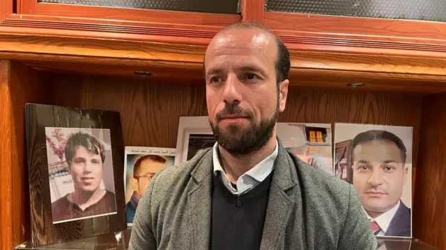 المحامي فادي العبار، ممثل رابطة عائلات قيصر في دمشق تظهر خلفه مجموعة صور.