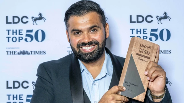 Manny lleva un elegante traje azul marino y camisa azul, sosteniendo a su izquierda un premio de los "LDC Top 50" y sonriendo a la cámara. Tiene el pelo oscuro y sonríe a la cámara con un anuncio promocional de LDC y The Times en un tablero detrás de él.
