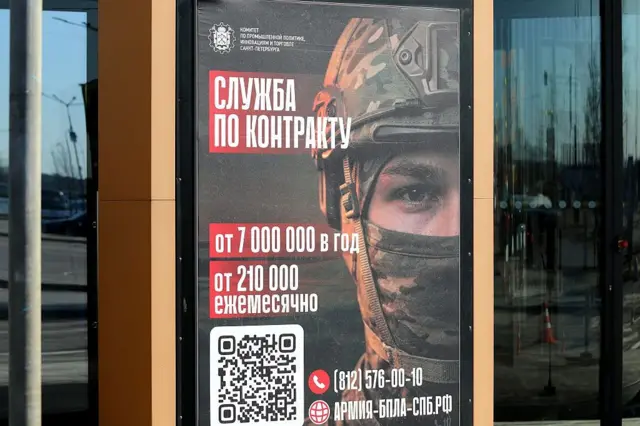 St. Petersburg'da asılı bir asker alım posterinde, miğfer takmış ve ağzı kapalı bir askerin başı görülüyor. Kırmızı bir zemin üzerinde, beyaz Rusça kelimeler, rakamlar, bir telefon numarası ve QR kodu, sözleşmeli askerlere yıllık yedi milyon rubleye kadar (aylık 210.000 ruble) maaş vaat ediyor. Posteri çevreleyen siyah camda ise bir sokak manzarası yansıyor.

