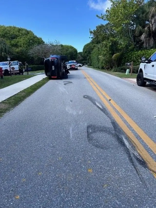 片側一車線の路上で黒い大型乗用車が横倒しになっている。路面にはタイヤ痕が見える。道の両側には芝生と歩道、木々が並ぶ
