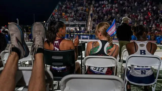 Три женщины сидят спиной к камере на переносных стульях на спортивном мероприятии. У одной из них — в центре — небольшой флаг трансгендеров.