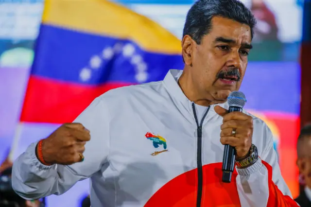 Maduro gesticulando enquanto fala ao microfone, com bandeira da Venezuela ao fundo 