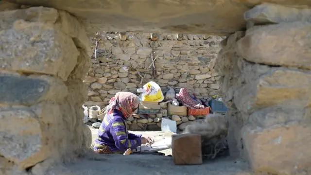A woman is seen through an opening in a stone wall in the foreground. She wears a headscarf and dress made of colourful fabrics and is sitting on the ground weaving wool. There are household belongings and another stone wall in the background. 