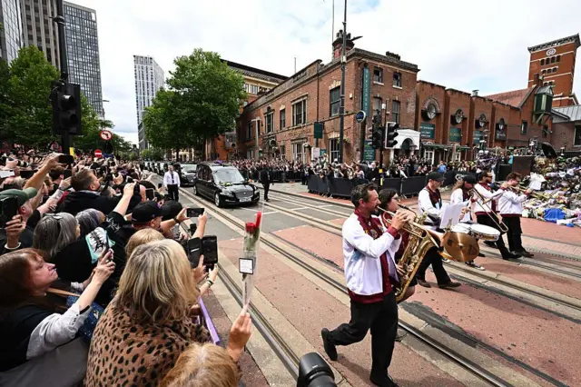 По трамвайным путям идет духовой оркестр, за ним движется кортеж из черных машин