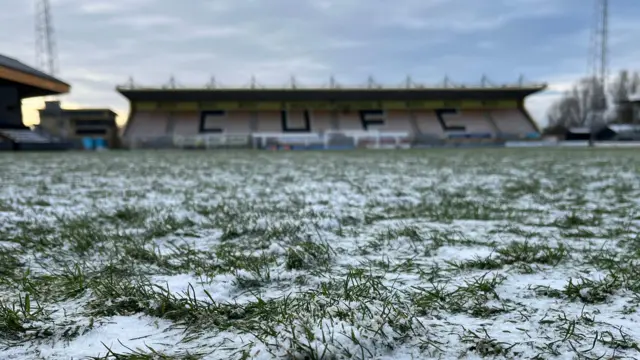 El césped de un campo de fútbol está cubierto por una fina capa de nieve y escarcha. Al fondo se ve una tribuna del estadio con el logo del COFC.