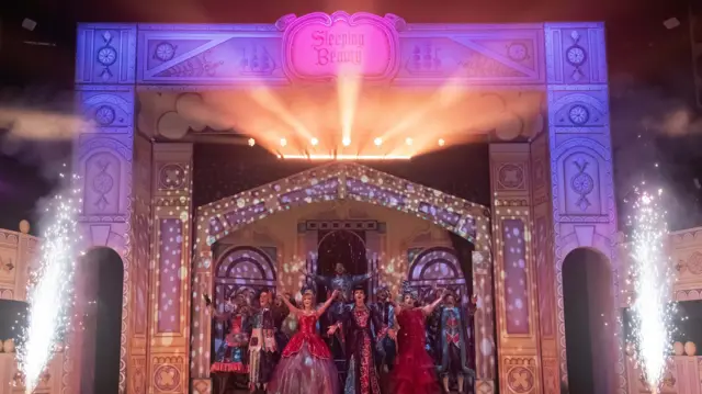 Actores con elaborados trajes actúan en un escenario. El escenario tiene un arco con un cartel que dice "La Bella Durmiente" en el centro. También hay una fachada de un edificio detrás de los actores.