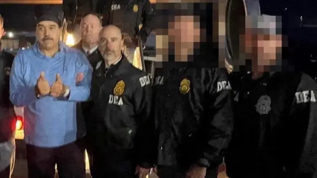 手錠をかけられたマドゥロ大統領と、その腕をとっているDEA長官がカメラに向かっている。他にDEA捜査員が周りに立ち、その多くが顔にモザイクがかけられている。マドゥロ氏は青いトップスに黒いズボン。頭に帽子かフードのようなものがのっている