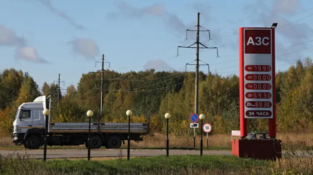 Дневной кадр у проселочной дороги в Нижегородской области: красная стела «АЗС» с ценами: АИ-95 - 79.99; АИ-92 и АИ-76 - 00.00; дизель и зимнее дизельное - 69.30; ниже указано «24 ЧАСА». Слева стоит грузовик, рядом дорожные знаки «остановка запрещена» для грузовиков и ограничение 10 км/ч; вокруг осенний лес и опоры ЛЭП.