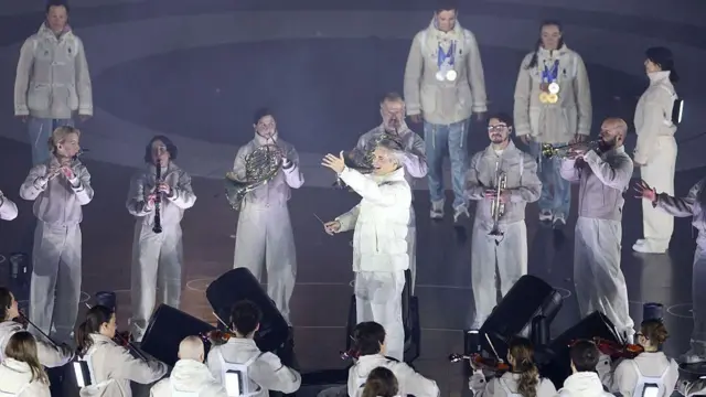 白っぽい衣装の人たちがさまざまな楽器を手に演奏している。中央では白っぽい衣装の指揮者が右手に指揮棒をもち、左手を開いて上方に伸ばして、口を開いている。背後にはメダルを首にかけた人たちが見える