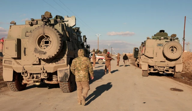 İki ağır zırhlı askeri aracın arasında 4 silahlı Suriye askeri var. Yol kapatılmış durumda. Hava açık ama soğuk olduğu giysilerden anlaşılıyor.