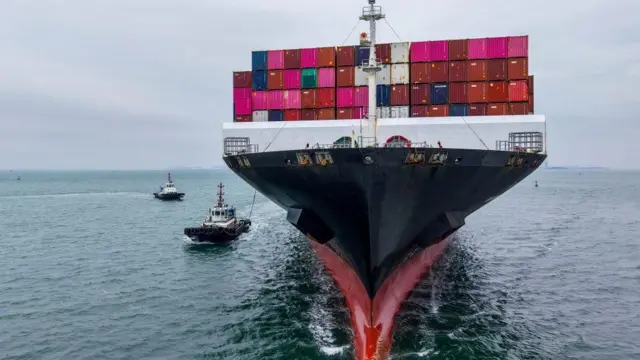 一艘大型貨輪正駛離鏡頭，船上堆滿了色彩鮮豔的貨櫃，包括亮粉紅、紅色、綠色和藍色。