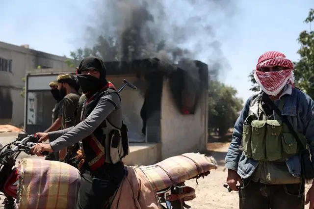 Бедуины на фоне дымящегося здания в деревне аль-Мазраа на юге Сирии