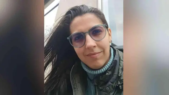 Vitória está olhando diretamente para a câmera. Ela veste uma blusa de gola alta azul, um casaco cáqui ou escuro, usa óculos e tem cabelos castanhos.