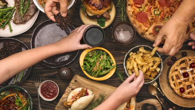 Mãos de diversas pessoas pegam alimentos, como batatas fritas, pizza, saladas e carne, de tigelas sobre uma mesa de madeira