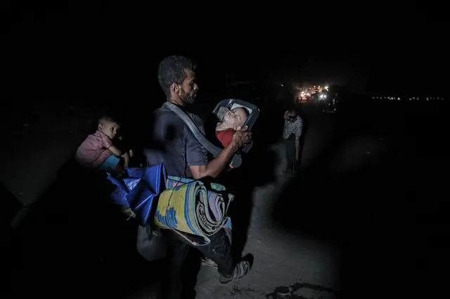 Man fleeing with his children on foot. He carries a sleeping child in a car chair slung across his shoulders, and has a carpet tied around his back