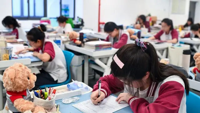 課室內,一名青少年坐在書桌前低頭看著桌上的紙張,並用黑色筆在紙上書寫。書桌上放有文具和一個玩具熊。背景可見多名青少年分散坐在各自的書桌前,身處同一課室環境。