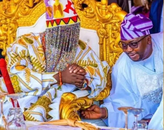 Alaafin Oyo joko lori itẹ, o de ade sori, wọ asọ oke funfun ati alawọ ofeefee, o n bọ minisita fun ohun amusagbara, Oloye Bayo Adelabu lọwọ, ẹni to wọn asọ agbada ati sokoto alawọ buluu, to si de fila alawọ elese aluko.