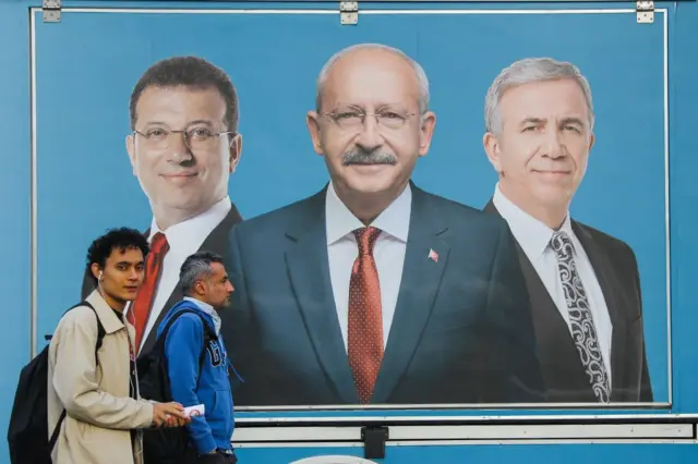 Ekrem İmamoğlu, Mansur Yavaş ve Kılıçdaroğlu fotoğrafları bir mavi fondalar. Bu bilboardun önünden insanlar geçiyor.