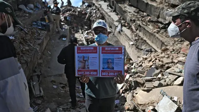 Hombre entre escombros mostrando las fotos de dos desaparecidos. 
