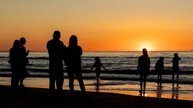 Sylwetki ludzi stojących na plaży i dzieci bawiących się w wodzie podczas zachodu słońca nad Morzem Bałtyckim 