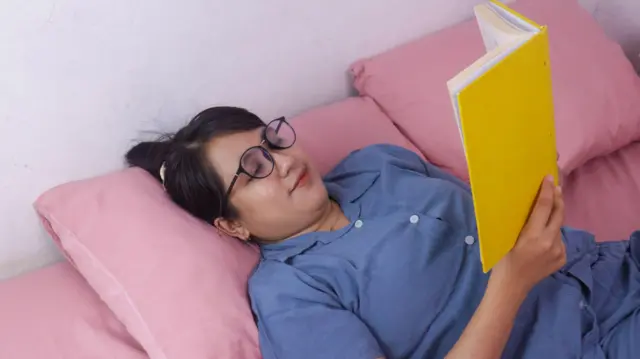 A young woman in pyjamas lies in bed, reading a book. She wears a pair of glasses and her hair is tied up. 