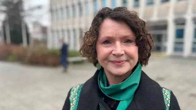 Tess Read lleva una bufanda de color jade y un abrigo de lana azul marino con detalles verdes y está parada frente a las oficinas del Ayuntamiento de Exeter, que están borrosas.