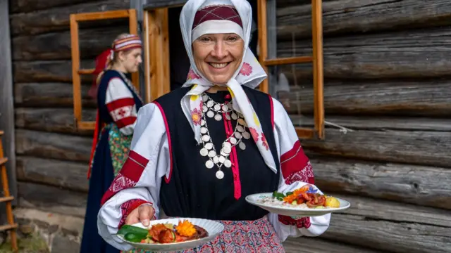 Hagyományos észt népviseletbe öltözött, ékszereket viselő nő két tányér ételt tart a kezében és mosolyog a kamerába. A háttérben faház látszik.