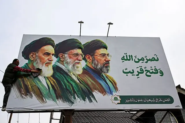 Um homem limpa um cartaz com as imagens dos falecidos líderes supremos do Irã, o aiatolá Ruhollah Khomeini (à esquerda) e o aiatolá Ali Khamenei (ao centro), ao lado do recém-eleito líder supremo, o aiatolá Mojtaba Khamenei