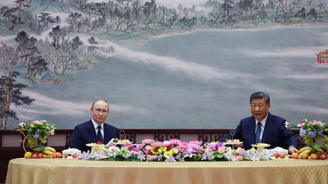 Путин и Си сидят за большим круглым столом с цветами. На стене за ними изображен зимний китайский пейзаж.