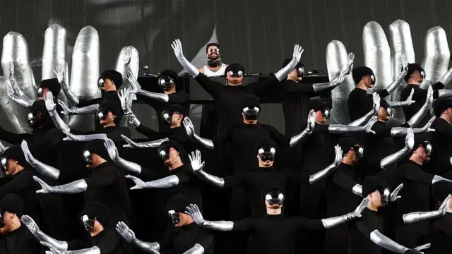 Alok se apresenta ao fundo do palco, cercado por dezenas de performers vestidos de preto, com braços prateados e máscaras espelhadas que ocultam seus rostos. Os dançarinos executam uma coreografia sincronizada com os braços erguidos em diferentes direções.