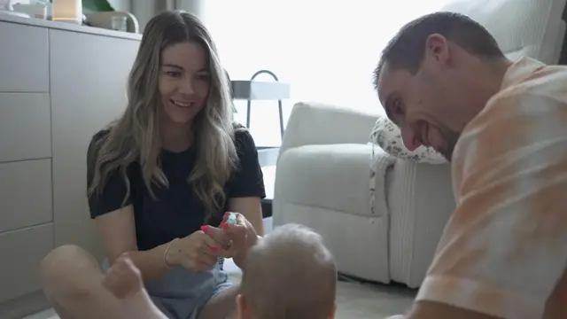 Egy hosszú szőke hajú nő és egy rövid barna hajú férfi a földön ülve játszanak a kisbabájukkal, akit hátulról látunk. A háttérben egy fotel.