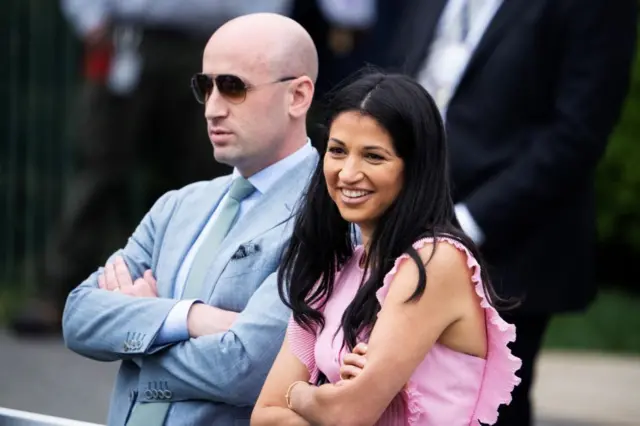 Katie Miller usa um vestido rosa e sorri. Ao seu lado, Stephen Miller veste um terno azul-claro e usa óculos escuros