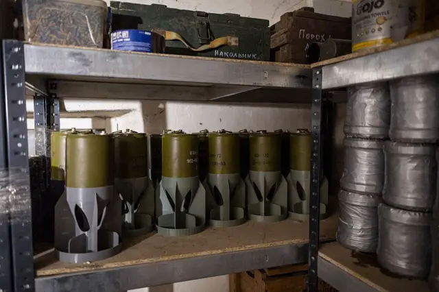 Металевий стелаж із декількома полицями, на яких в ряд стоять зелені циліндричні боєприпаси з металевими хвостовими стабілізаторами. Праворуч на іншій полиці розміщені кілька великих рулонів, щільно обмотаних сріблястою ізоляційною стрічкою.