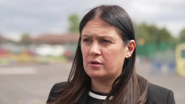 暗い色の長髪で、黒いスーツを着た女性がインタビューに答えている