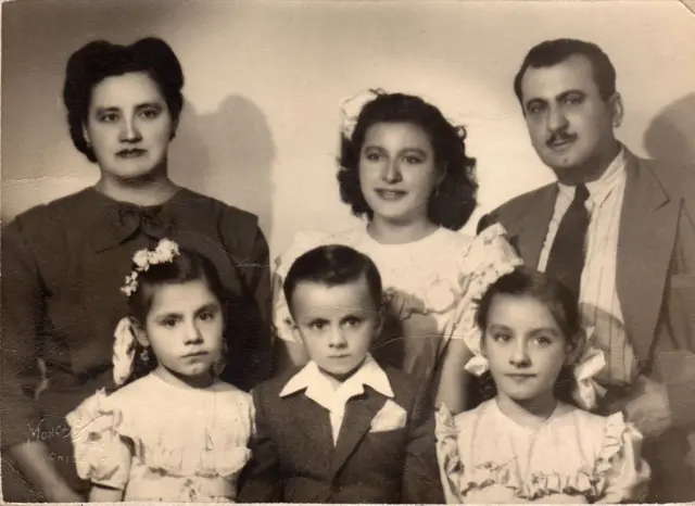La madre de Alejandra Jáidar, con un vestido oscuro, está parada en la parte izquierda. A su lado y en el centro de la foto está Alejandra, de adolescente, con un vestido blanco y un adorno blanco en la cabeza. A su lado está su padre, vestido de traje y corbata. Delante de ellos están tres niños, uno de ellos es un niño que está en el centro