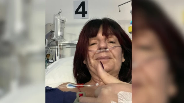 Trish has a fringe and short hair. She is lying in a hospital bed with her thumb up. There is a tube coming out of her nose and she is wearing a dark blue top. Behind her an IV drip can be seen.