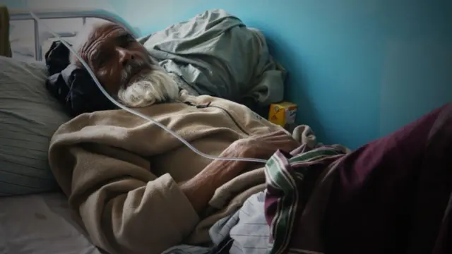 ایک بوڑھا شخص ہسپتال کے بستر پر لیٹا ہے، اُس کے بازو میں ڈرِپ لگی ہوئی ہے اور وہ ایک نیم تاریک کمرے میں کیمرے کی طرف دیکھ رہا ہے