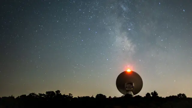 Gece vakti bir teleskobun fotoğrafı; iki yanında ağaçların siluetleri görülüyor; yukarıdaki yıldızlı gece gökyüzünde Samanyolu galaksisi seçilebiliyor.