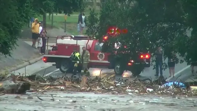 手前に氾濫した川と、川が運んだがれきが積み上がり、その奥で消防服を着た人や住民らが様子を見ている。緊急対応当局の赤い車両も停まっている