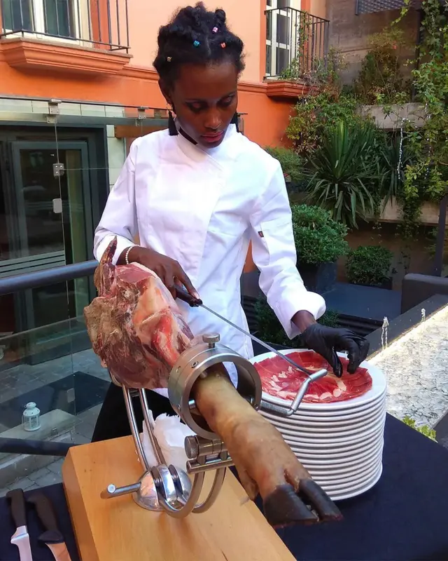Afroamerikanka reže komade šunke u kuvarskoj uniformi