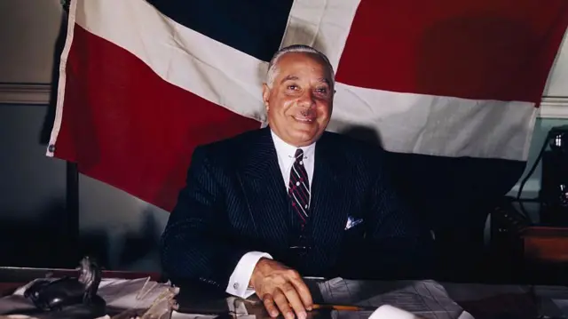 Rafael L. M. Trujillo, presidente de la República Dominicana, sonriendo con una bandera del país detrás.