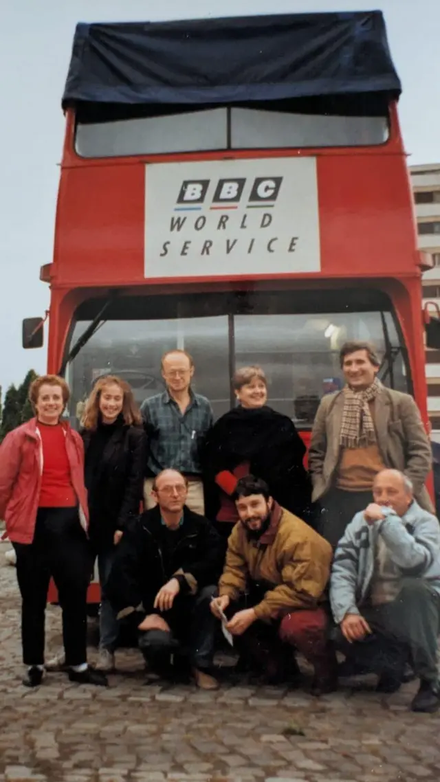Opt persoane, femei și bărbați, stau pentru o fotografie, zâmbind, în fața unui autobuz roșu supraetajat pe care scrie BBC World Service.