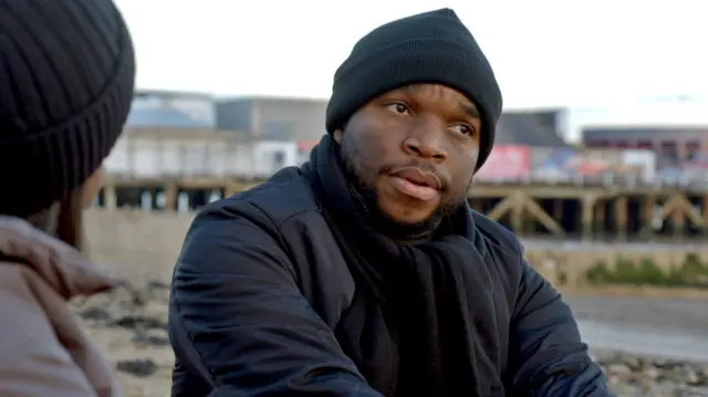 Praise na Nigerian man wey dey im mid 30s, wey wear black beanie, navy coat and black scarf for di sea front in Clacton-on-Sea.