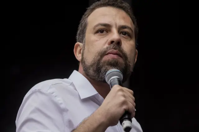 Foto em close do rosto de Guilherme Boulos, com semblante sério. Ele segura microfone em um evento de campanha.